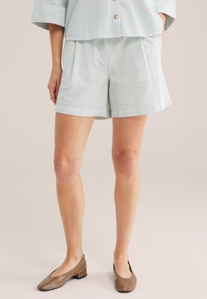 Žena oblečená v svetlosivých prúžkovaných šortkách v kombinácii so zodpovedajúcim topom a hnedými balerínami, stojaca s jednou rukou vo vrecku.