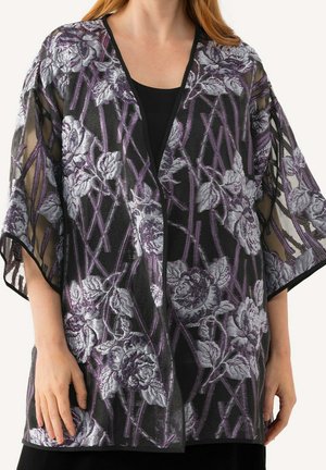 Elle porte un kimono noir transparent avec une broderie florale violette et grise sur un haut noir, les mains détendues le long du corps.