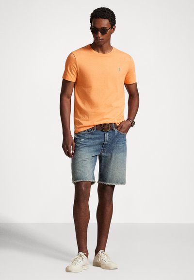 Φωτεινό πορτοκαλί βαμβακερό μπλουζάκι με στρογγυλή λαιμόκοψη, φορεμένο με ξεθωριασμένα τζιν σορτς και ανοιχτόχρωμα αθλητικά παπούτσια. Τα αξεσουάρ περιλαμβάνουν γυαλιά ηλίου και ζώνη με υφή.