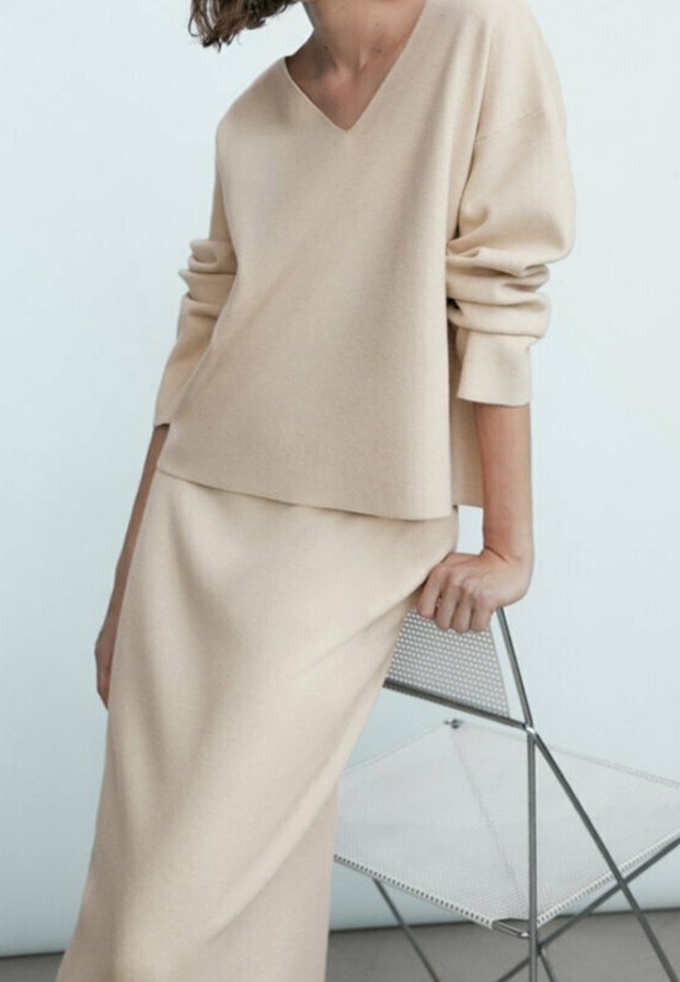 Maglione a maglia beige con scollo a V e maniche ampie, abbinato a una gonna lunga fino a terra dello stesso colore. Texture liscia e design minimale.