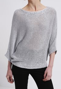 Hellgrauer, oversized Strickpullover mit lockerem Gewebe, weitem Ausschnitt und Dreiviertelärmeln. Kombiniert mit schwarzen, eng anliegenden Hosen.