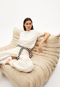 Beige, pluche bank met een gequilt ontwerp en een zachte textuur. Het model draagt een witte top en crèmekleurige broek, afgewerkt met een zwart-wit gestreepte riem.