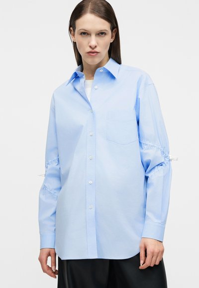 Γυναίκα που φοράει μια ανοιχτόχρωμη μπλε πουκαμίσα με κουμπιά, φαρδιά εφαρμογή, με ραβδώσεις και κορδόνια στα μανίκια και τσέπη στο στήθος, συνδυασμένη με μαύρο παντελόνι.