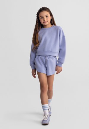 Niña con cabello largo que lleva una sudadera color lavanda, pantalones cortos a juego, calcetines a rayas y zapatillas deportivas, de pie frente a un fondo liso.