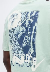 T-shirt vert clair avec un graphisme bleu et blanc d’un joueur de basketball en train de dunker et le texte « Nike » et « The History ».