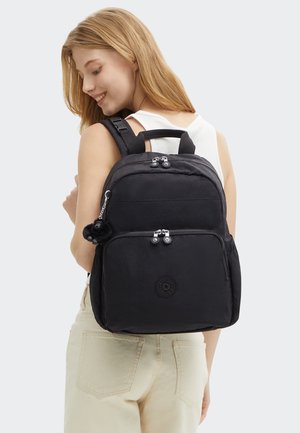 Giovane donna con capelli biondi che indossa una canottiera bianca e pantaloni beige, porta uno zaino nero con tasche con cerniera e un piccolo portachiavi di peluche.