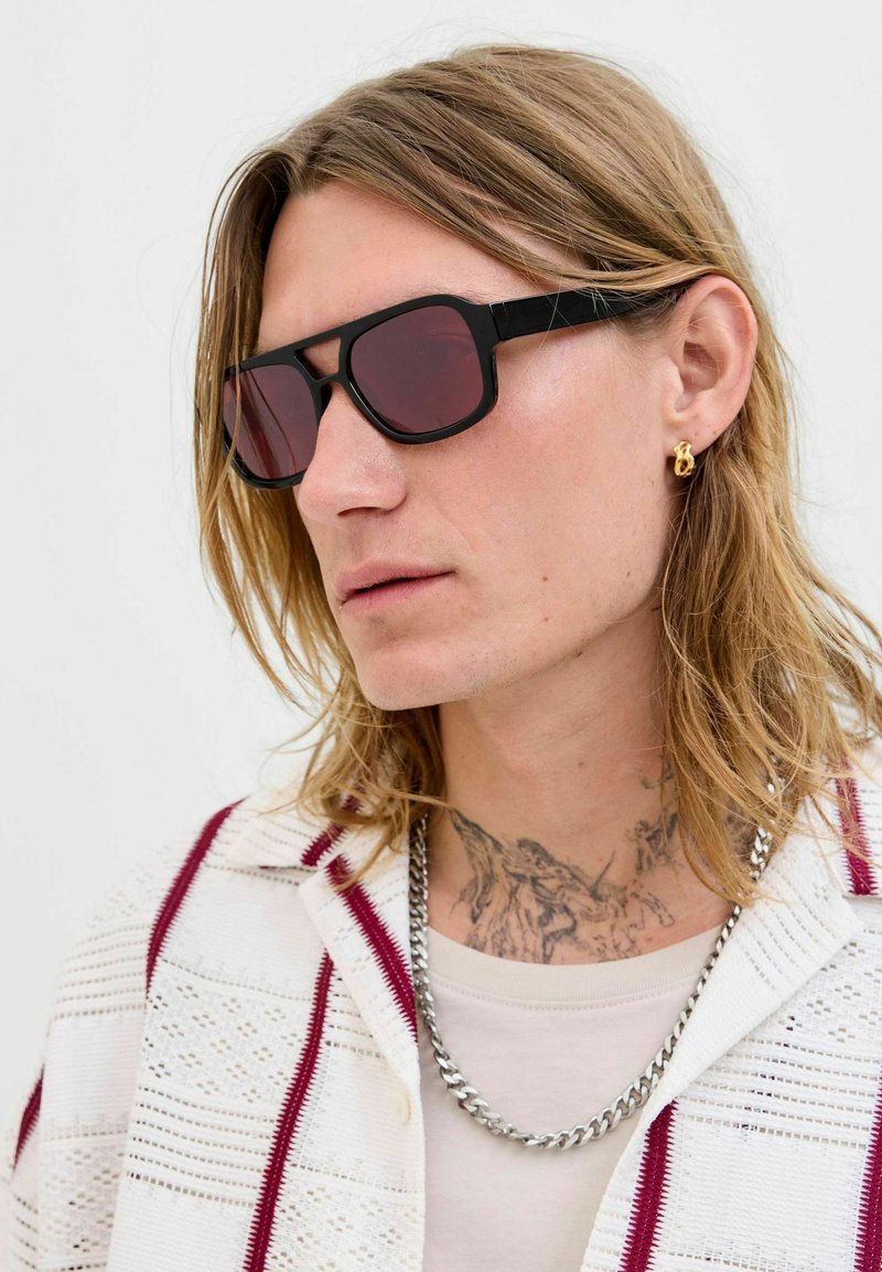 Schwarze Sonnenbrille mit rechteckigen Rahmen und kastanienbraun getönten Gläsern. Getragen mit einem weißen gemusterten Hemd und einer silbernen Kettenhalskette.