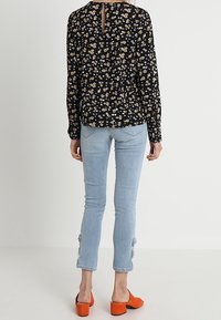 Blouse noire à motif floral avec manches longues, dos échancré, associée à un jean skinny bleu clair avec des détails de boutons à l'ourlet et des talons orange.