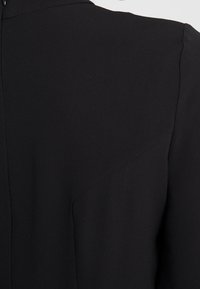 Czarna odzież o fakturowanej powierzchni, z wysokim dekoltem i kątowymi szwami na ramionach. Materiał wydaje się gładki i lekki.