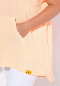 Tunic di un arancione chiaro, con texture, dotata di una tasca frontale e un'etichetta gialla sull'orlo. Abbinato a pantaloni bianchi, che evidenziano un design morbido.