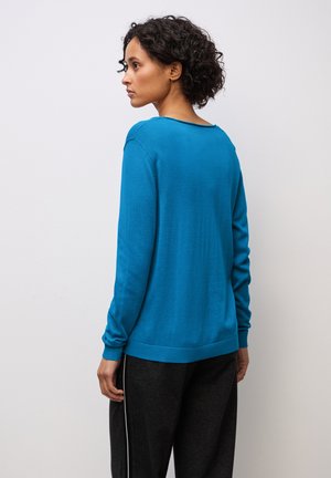 Pull en tricot bleu avec une coupe décontractée, des poignets et un ourlet côtelés, et un large décolleté. Porté avec un pantalon de jogging gris foncé, affichant un style décontracté.