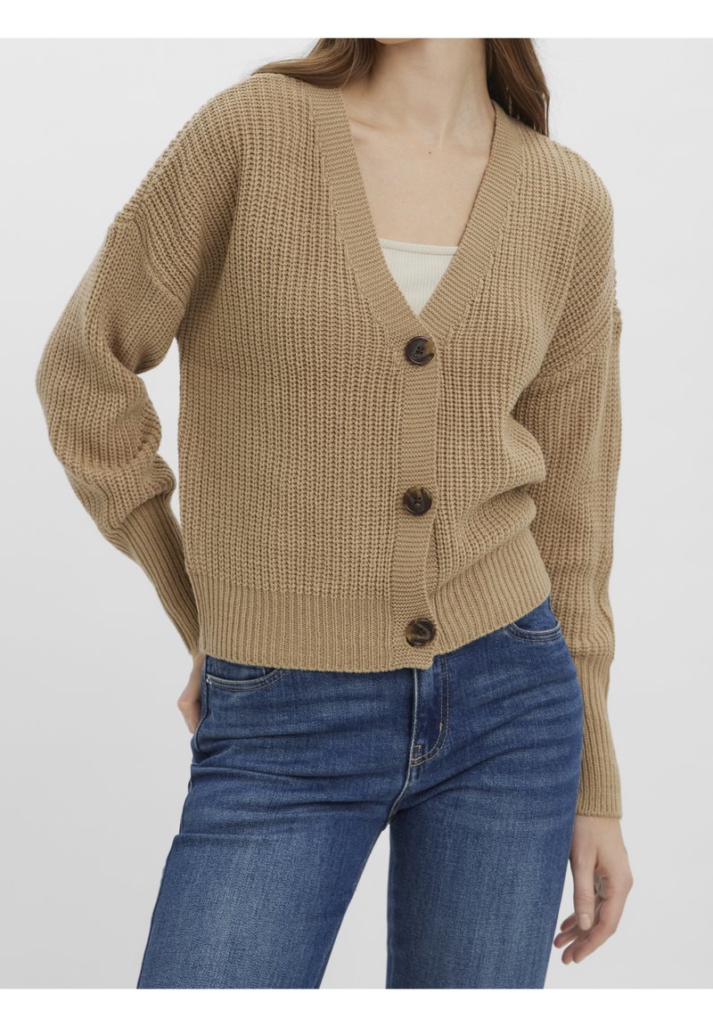 Femme portant un cardigan beige en maille boutonné par-dessus un haut blanc et un jean bleu, debout avec une main sur la hanche devant un fond uni.