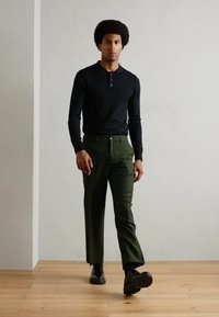 Chemise henley noire à manches longues avec boutons, assortie à un pantalon ajusté vert olive et des chaussures noires, sur fond gris clair.