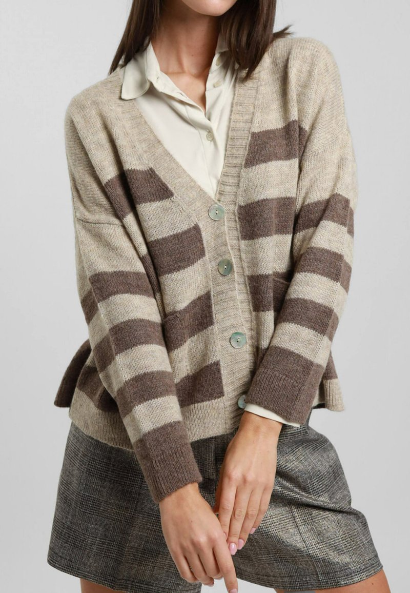 Cardigan lavorato a maglia beige con strisce marroni, scollatura a V, bottoni anteriori e spacchi laterali. Indossato sopra una blusa chiara e una gonna texturizzata.