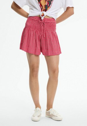 Persona que lleva pantalones cortos a cuadros rojos y blancos con cintura fruncida y zapatillas blancas, de pie con las manos en las caderas.