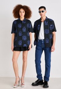 Zwarte T-shirts met korte mouwen hebben blauwe grafische prints. Het vrouwelijke model draagt een zwarte rok en zilveren hakken, terwijl het mannelijke model blauwe jeans en zwarte schoenen heeft.