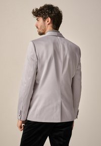 Hellgrauer, maßgeschneiderter Blazer mit glatter Textur, einem Schlitzausschnitt hinten und glänzenden Knöpfen an den Ärmeln, kombiniert mit dunklen Hosen.