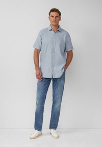 Camicia a maniche corte, blu chiaro, con bottoni e un motivo texturizzato, abbinata a jeans in denim chiaro e sneakers bianche.