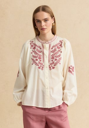 Jeune femme portant un chemisier crème avec broderies florales roses et un pantalon rose, debout devant un fond beige.