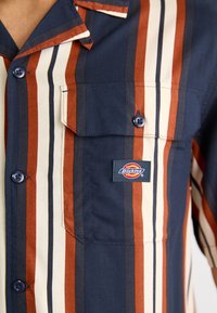 Cămașă cu mânecă scurtă, cu dungi verticale în nuanțe de bleumarin, arămie și crem, având două buzunare frontale și un logo Dickies brodat pe pieptul stâng.
