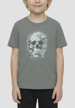 Bambino che indossa una maglietta grigia con un grande disegno di teschio circondato da foglie di felce sul davanti su uno sfondo semplice.