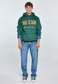 Jongeman in een groene "BIG STAR" hoodie, blauwe geruite shirt, lichtblauwe jeans en groene sneakers, staand tegen een effen achtergrond.