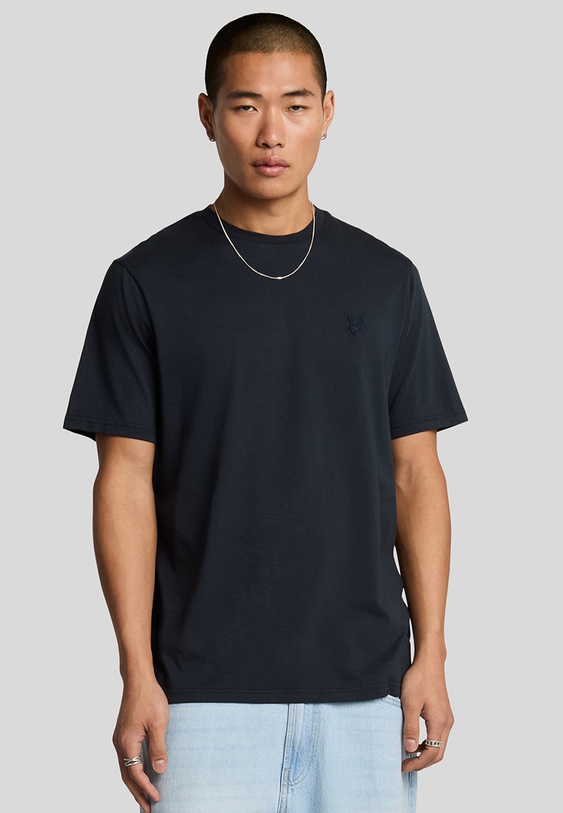 Camiseta de algodón negra de manga corta, con cuello redondo y logo bordado en el pecho, combinada con pantalones cortos de mezclilla azul claro.