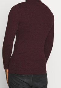 Person som bär en åtsittande, ribbad, mörk bourgognefärgad polotröja och svarta jeans, visad bakifrån mot en vit bakgrund.