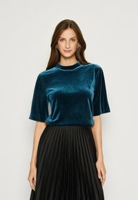 Teal fluwelen top met een hoge hals en korte mouwen, gecombineerd met een geplooide zwarte rok. De stof heeft een gladde, rijke textuur.