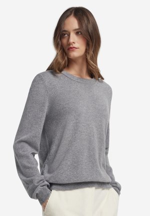Femme aux cheveux châtain mi-longs portant un pull gris clair à manches longues et un pantalon écru, les mains dans les poches.