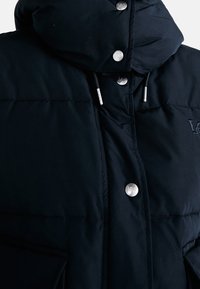 Svart puffjacka med hög krage, silverfärgade tryckknappar och dragsko. Tyget verkar jämnt och vadderat för isolering.