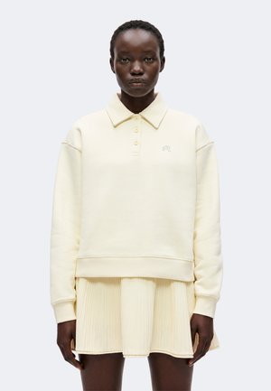 Kvinde iført en cremefarvet langærmet sweatshirt med krave og et matchende plisseret nederdel, stående mod en ensfarvet lys baggrund.