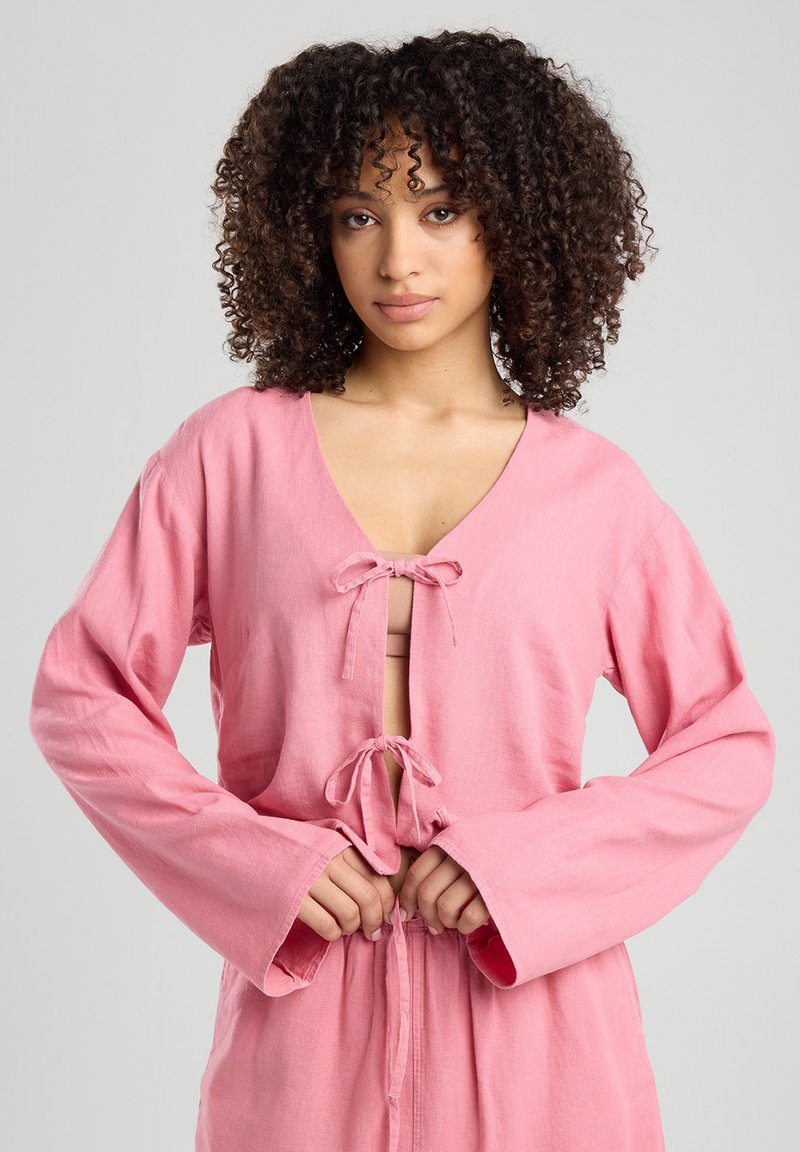 Indumento rosa a maniche lunghe con scollatura a V, caratterizzato da dettagli annodati al petto e in vita, realizzato in un tessuto texturizzato.