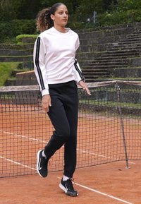 Λευκό φανελάκι με μαύρες ρίγες στα μανίκια, μαύρο παντελόνι φόρμας και μαύρα αθλητικά παπούτσια. Περπατά σε χωμάτινη τένις κοντά σε δίχτυ.