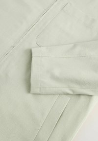 Tejido texturizado de color verde claro con una manga doblada y un bolsillo frontal, que presenta un diseño limpio y estructurado con detalles de costura visibles.