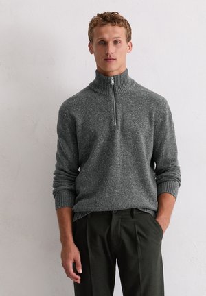 Grijze zip-up trui met ribgebreide kraag en manchetten, met een gevarieerd breipatroon. Gedragen over donkergroene broeken.