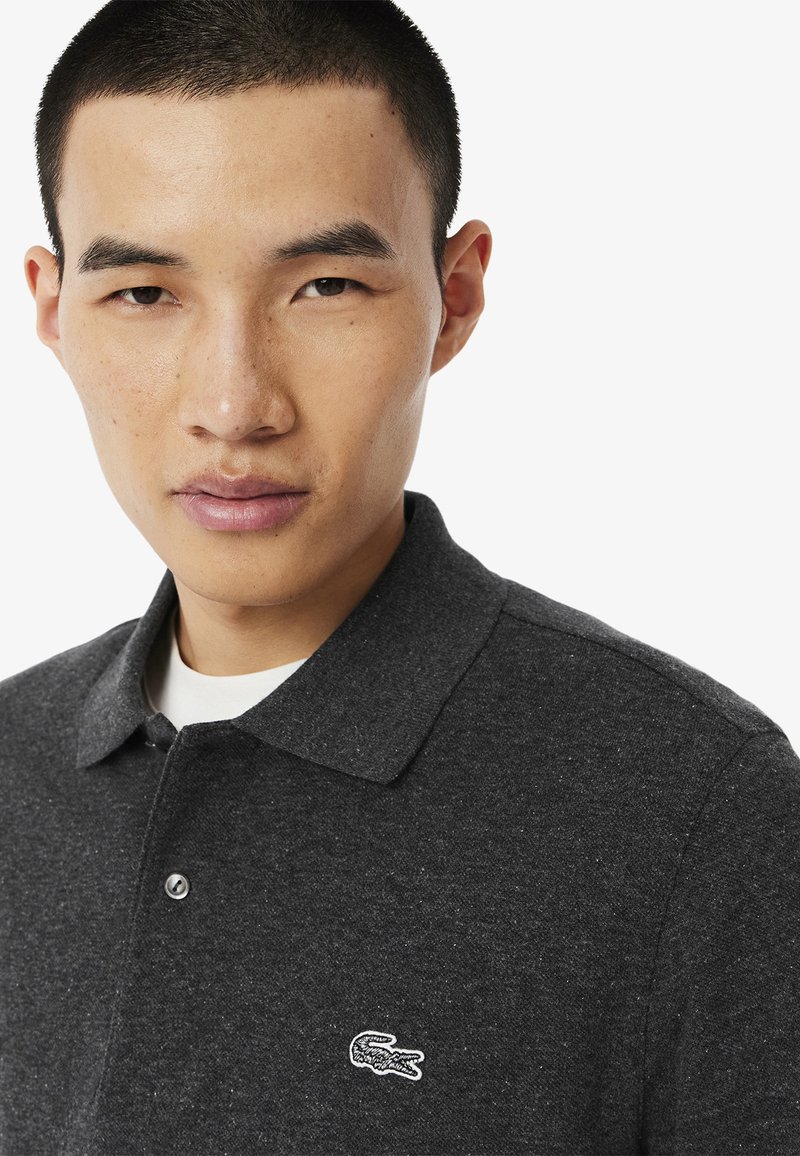 Polo shirt gris foncé en tissu texturé, avec un petit logo crocodile brodé blanc sur la poitrine gauche et un col boutonné.