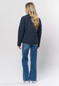 Vrouw met lang blond haar, gekleed in een zwart doorgestikt jack, blauwe spijkerbroek en witte sneakers, staand met haar rug naar de camera tegen een effen achtergrond.