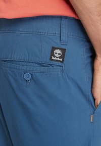 Blauwe katoenen shorts hebben een achterzak met een blauwe knoop en een zwart Timberland logo patch. De stof heeft een gladde textuur.