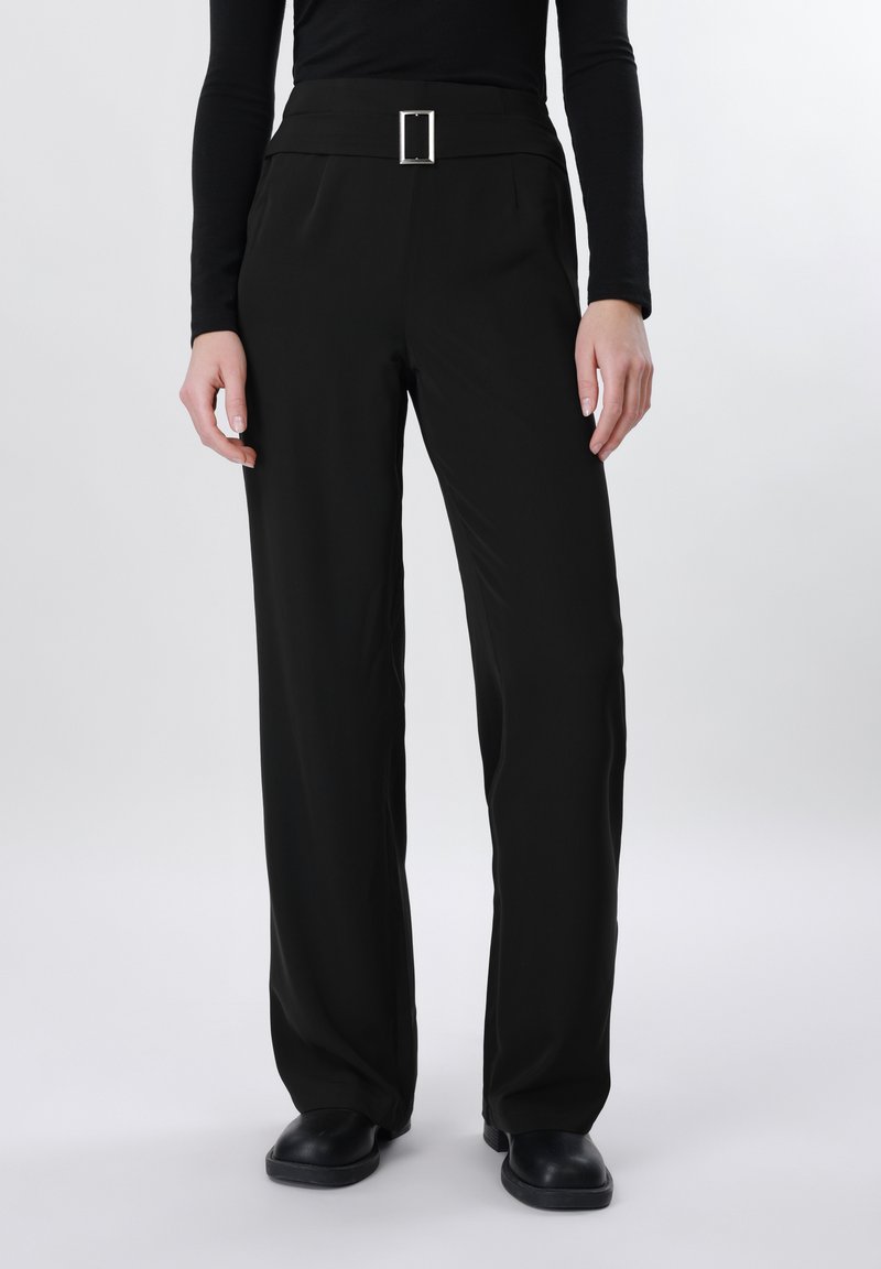 Άτομο που φοράει μαύρο παντελόνι με ψηλή μέση και φαρδιά μπατζάκια με τετράγωνη αγκράφα στη ζώνη, συνδυασμένο με μαύρα παπούτσια και μαύρο τοπ με μακριά μανίκια.