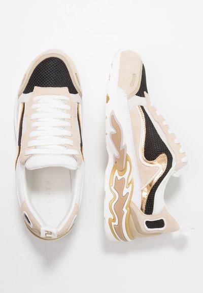 Beiga och svarta sneakers har ett ovanparti i mesh, guldaccenter och vita snören. Designen inkluderar en texturerad tå och en sula med vågmönster.