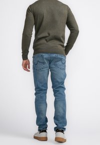 Outfit da uomo con un maglione verde oliva, jeans in denim blu chiaro con dettagli sulla tasca posteriore e sneaker grigie con suole marroni.