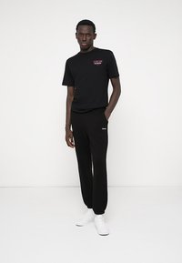 Melnas T-krekls ar drukātu logotipu, savienots ar melnām sporta biksēm. Abiem ir brīvs piegriezums; ansamblis tiek pabeigts ar baltām sporta apavām.