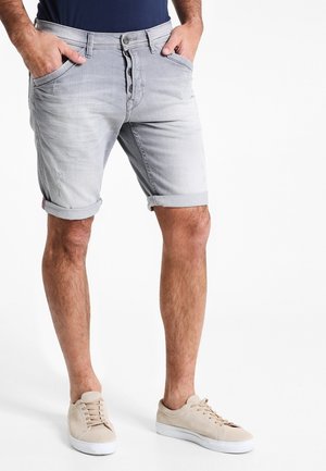 Mand iført lysegrå, opviklede denimshorts og beige sneakers, stående med hænderne i lommerne.