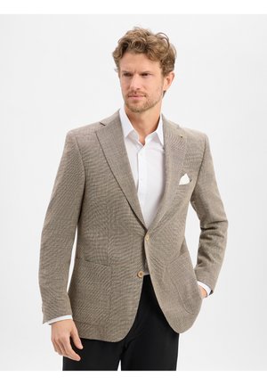 Mann in beigem strukturiertem Blazer, weißem Hemd und schwarzer Hose, stehend vor einem schlichten hellen Hintergrund, mit einer Hand in der Tasche.