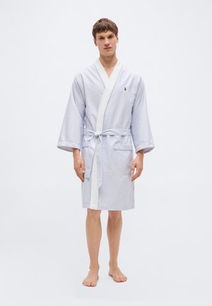 Jonge man staat op blote voeten en draagt een lichtblauw-wit gestreepte badjas tot op de knieën met een ceintuur in de taille.