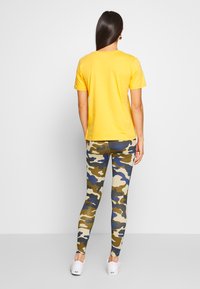Gul kortermet t-skjorte kombinert med flerfargede kamuflasje leggings med beige, grønne, blå og brune mønstre; brukt med hvite sko.