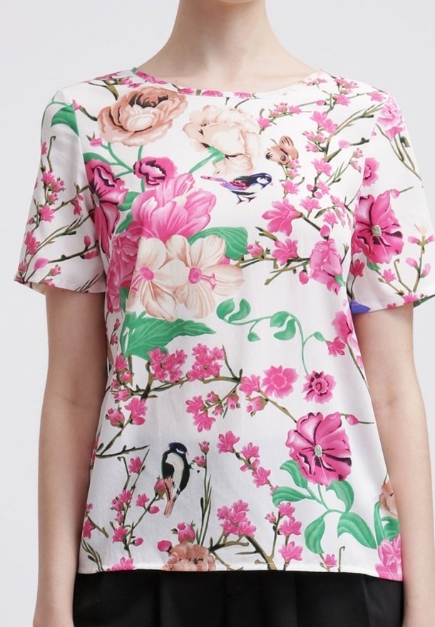 Chemise blanche à manches courtes avec des motifs floraux et d'oiseaux rose, beige et verts, portée par une personne des épaules aux hanches.
