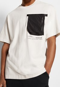 Krátký rukáv krémové tričko s černou zipovou kapsou, tištěný textový design na přední straně a volný střih. Textura z bavlněné směsi.