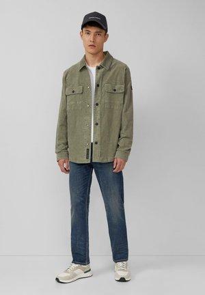 Olijfgroene corduroy jas met twee borstzakken, gecombineerd met een wit shirt, blauwe spijkerbroek en licht meerkleurige sneakers.